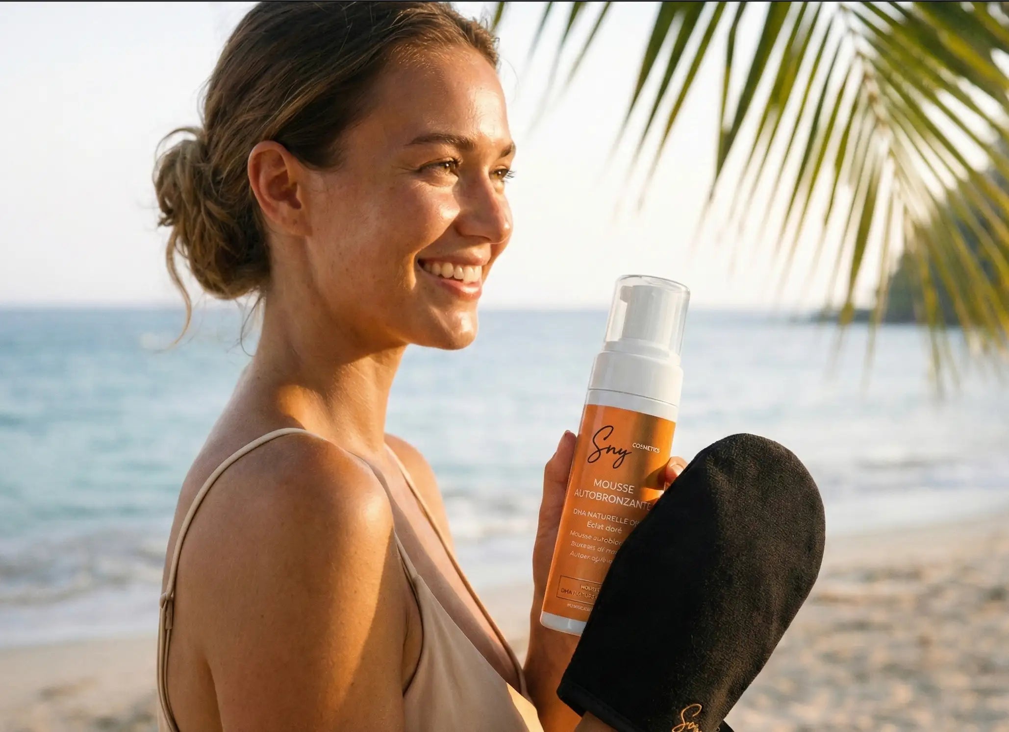 Femme souriante sur une plage au coucher du soleil tenant la mousse autobronzante SNY Cosmetics et portant le gant applicateur noir, éclairée par une lumière dorée naturelle.